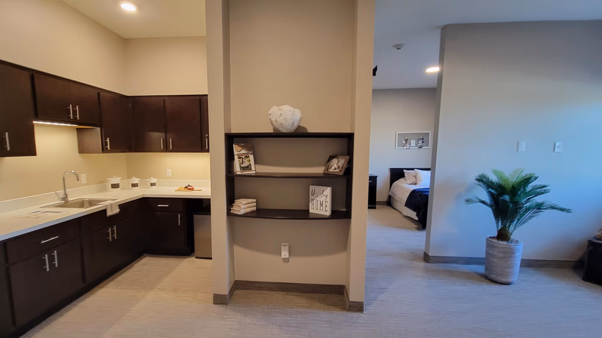 Interior view of a senior living facility showing a kitchen area with dark wood cabinets and a sink on the left, a small shelving unit in the center with decorative items, and a bedroom visible in the background on the right with a bed and framed pictures on the wall. A potted plant is placed near the bedroom entrance.