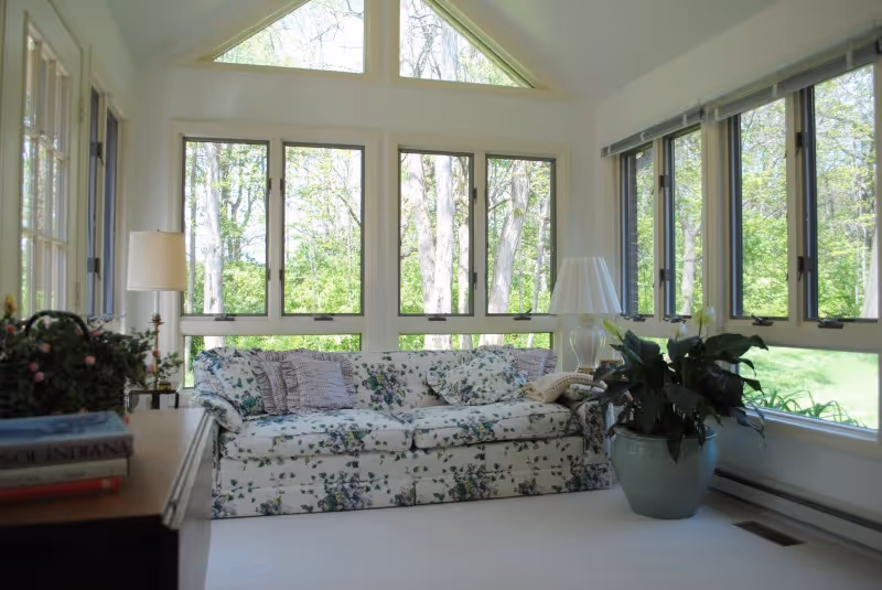 A bright living room with large windows showing a view of trees outside. The room features a floral-patterned sofa with several cushions, a large potted plant, two table lamps, and a wooden side table with books and a small plant on it.