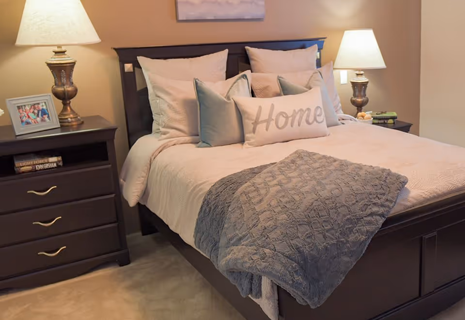 A neatly made bed with multiple pillows including one with the word 'Home' on it, a folded gray blanket at the foot of the bed, two bedside tables each with a lamp, a framed photo on the left table, and books on the shelves beneath the tables.