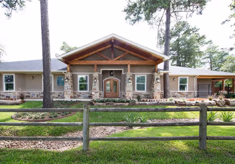 Front exterior view of Village Green Memory Care Community Tomball building with a wooden fence in the foreground, stone pillars supporting a wooden gabled roof over the entrance, surrounded by green grass, trees, and landscaping.