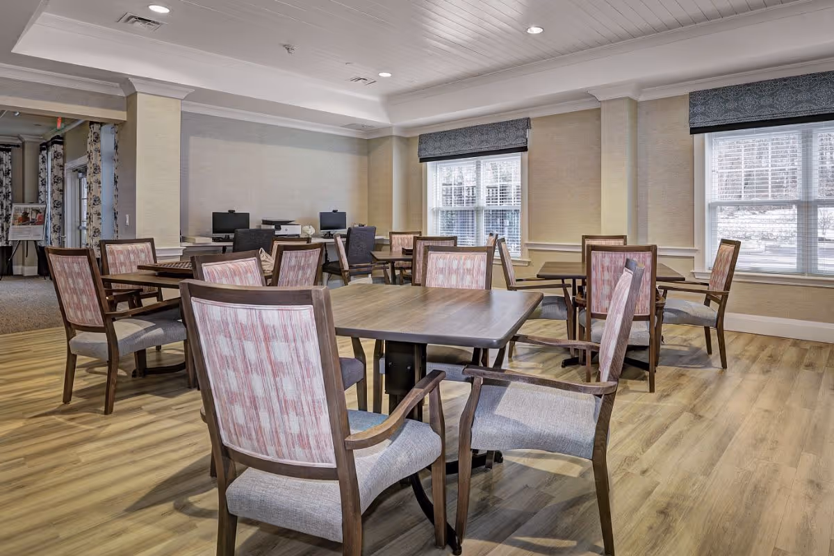 A bright room with several wooden tables and chairs with patterned upholstery arranged neatly on a light wood floor. The room has large windows with blinds and valances, allowing natural light to fill the space. In the background, there are two computer workstations with chairs. The walls are painted in a neutral tone with white trim and crown molding.