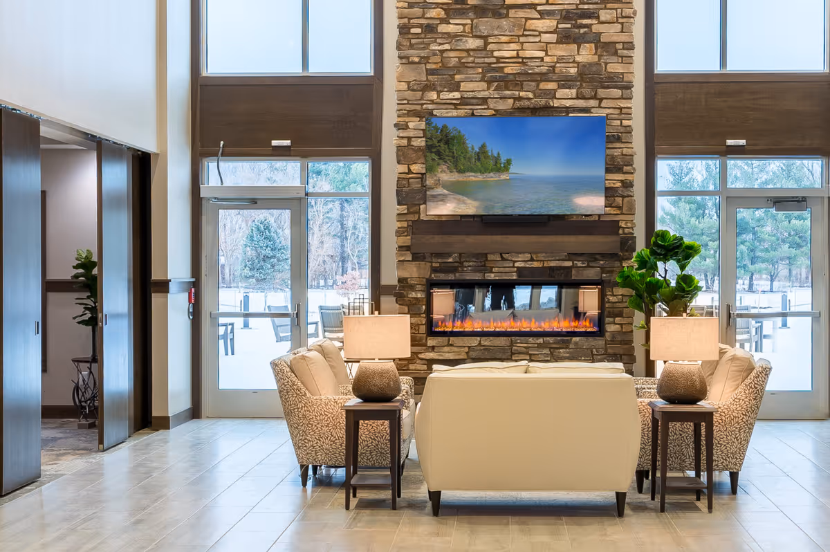 A cozy seating area in a senior living facility with a stone fireplace and a flat-screen TV mounted above it. The seating includes a white sofa and two patterned armchairs, each with a side table and lamp. Large windows and glass doors behind the seating area provide a view of an outdoor patio with trees.
