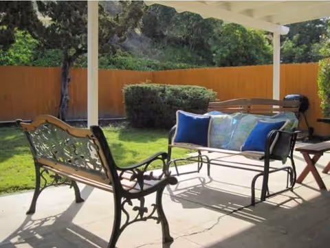 Outdoor patio area with two metal benches, one with blue and patterned cushions, a small wooden side table, and a barbecue grill. The patio is covered by a white pergola and overlooks a green lawn with bushes and trees, enclosed by a wooden fence.
