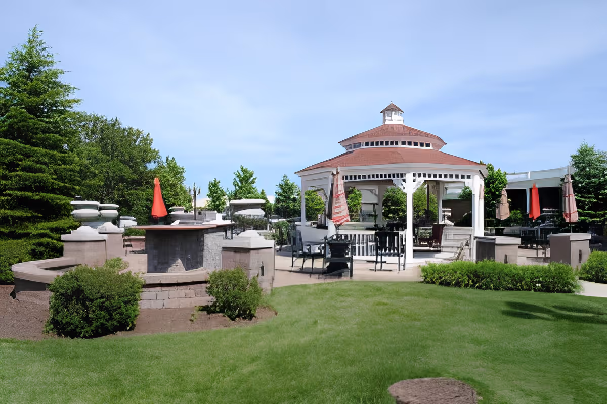 A landscaped outdoor patio featuring a white gazebo surrounded by tables, chairs, red umbrellas, and green lawn.