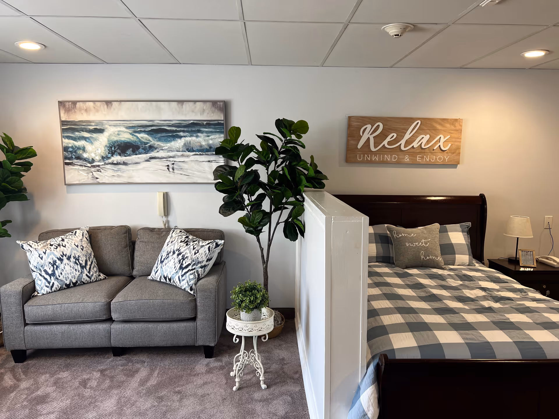 A cozy living space featuring a gray loveseat with patterned pillows on the left, a large potted plant in the center, and a bed with a checkered comforter and pillows on the right. Above the loveseat is a painting of ocean waves, and above the bed is a wooden sign that reads 'Relax Unwind & Enjoy'. A small white decorative table with a plant sits next to the bed, and a nightstand with a lamp and phone is visible beside the bed.