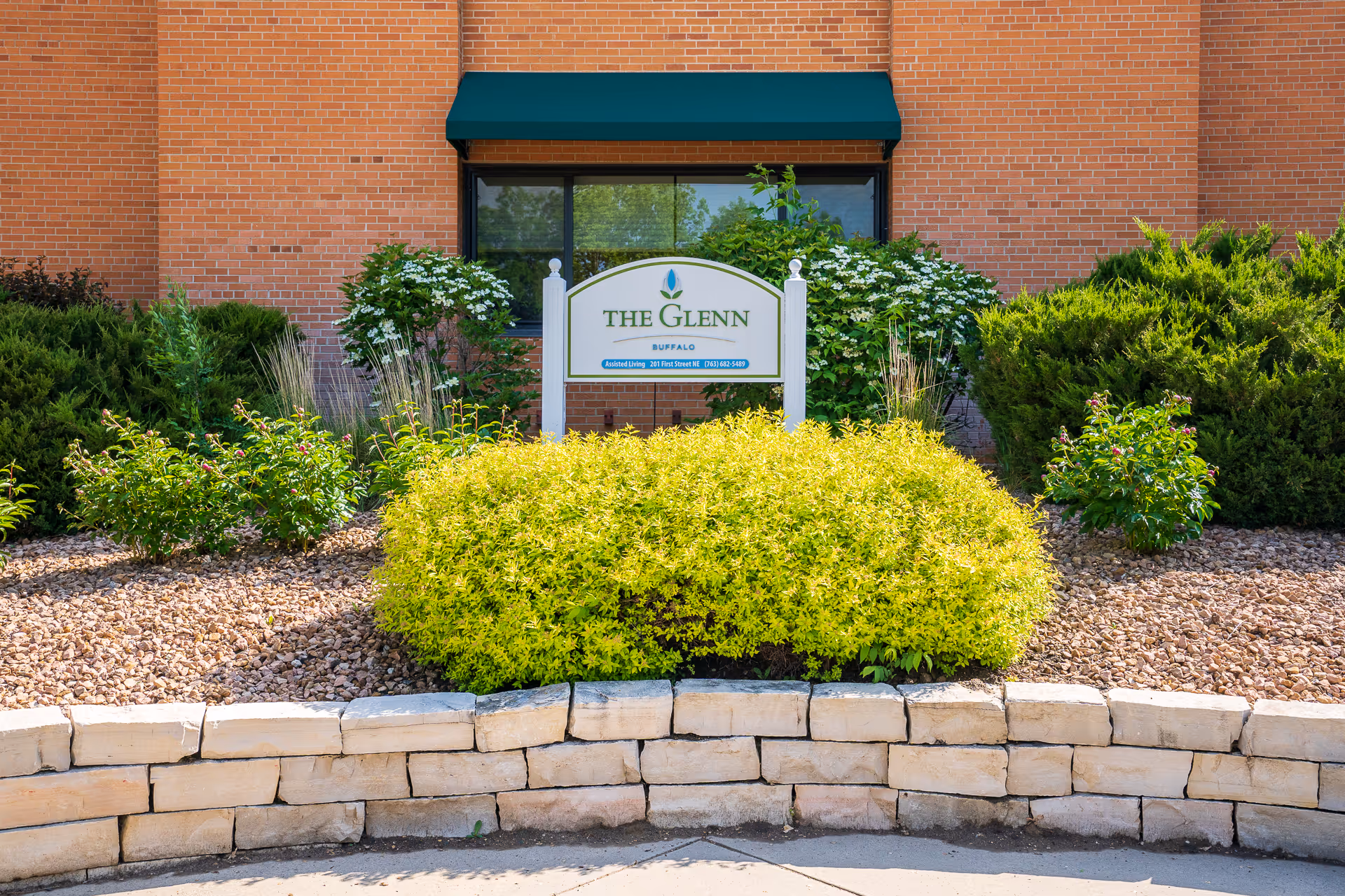 Outdoor view of the entrance area of The Glenn Buffalo facility, featuring a landscaped garden with green shrubs and plants, a stone retaining wall, and a sign with the facility's name in front of a brick building with a window and green awning.