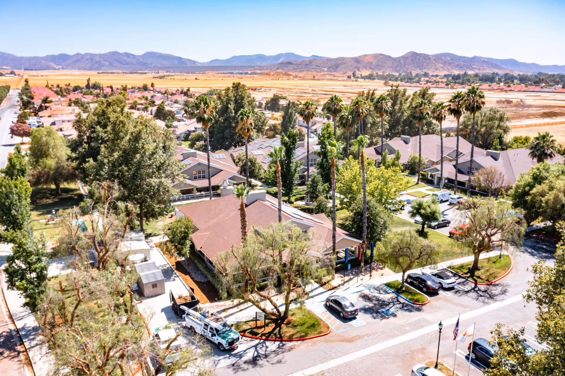 Aerial view of a senior living facility named Cottages at Hemet, showing multiple buildings with brown roofs surrounded by tall palm trees and other greenery. There are several parked cars in the parking lot, and mountains are visible in the background under a clear blue sky.