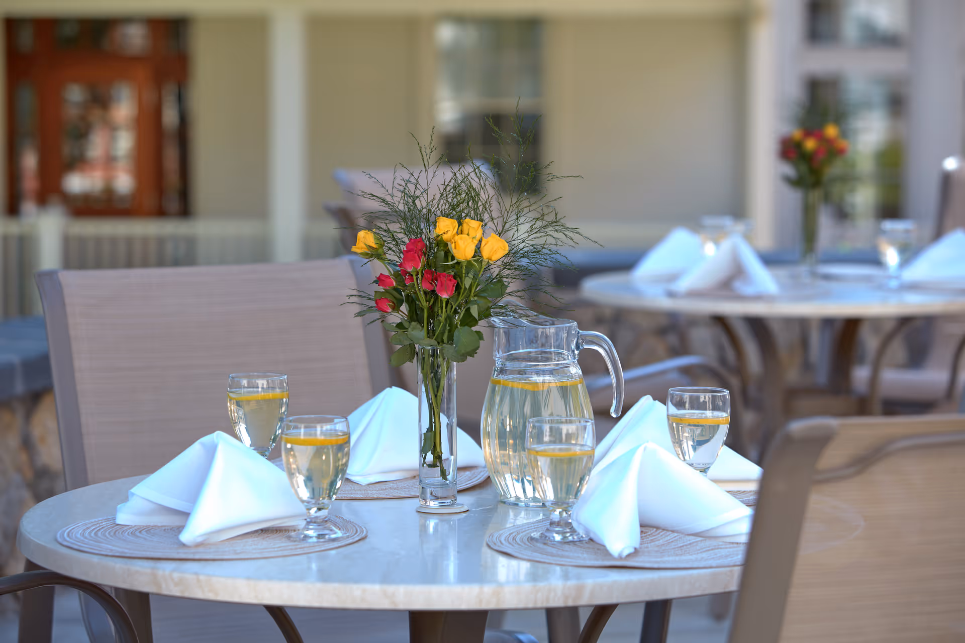 Outdoor patio dining table set with a vase of yellow and red roses, a pitcher of water with lemon, glasses, and folded napkins.