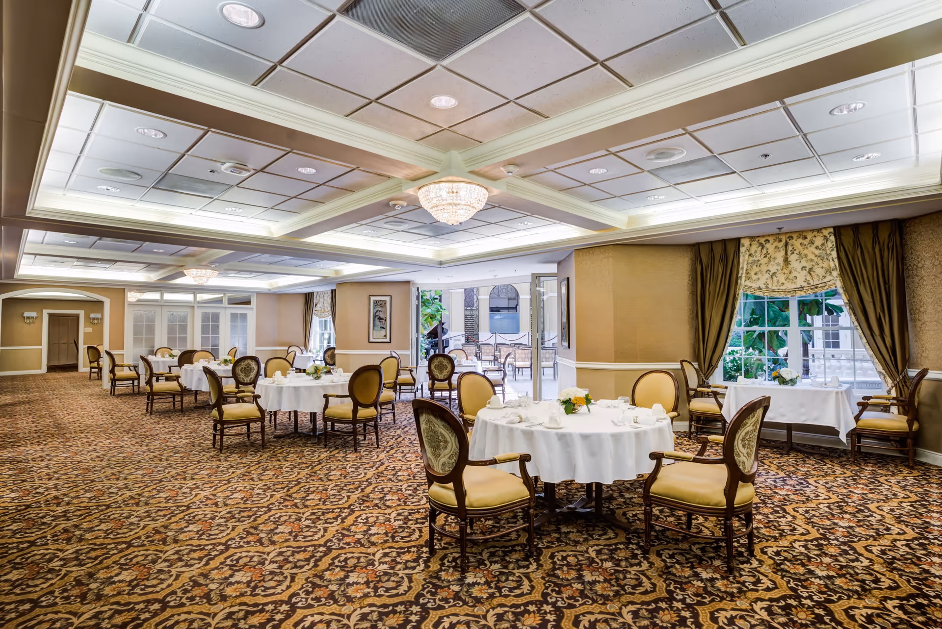 Elegant dining room with round tables covered in white tablecloths, surrounded by upholstered chairs. The room features patterned carpet, a coffered ceiling with recessed lighting and chandeliers, and large windows with drapes allowing natural light. There is an open doorway leading to an outdoor patio area with additional seating.
