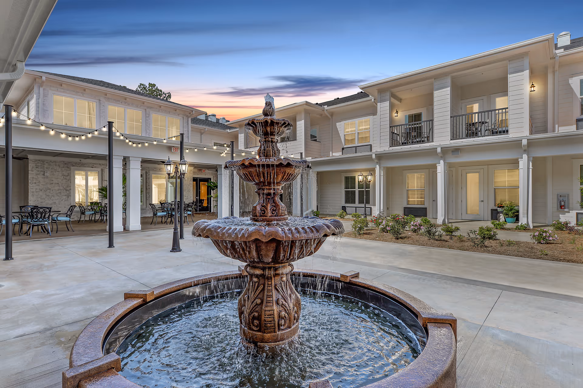 Outdoor courtyard area at The Blake at Biloxi featuring a large three-tiered water fountain in the center, surrounded by a paved patio with string lights overhead, outdoor seating with tables and chairs, and a two-story building with balconies and lit windows at dusk.