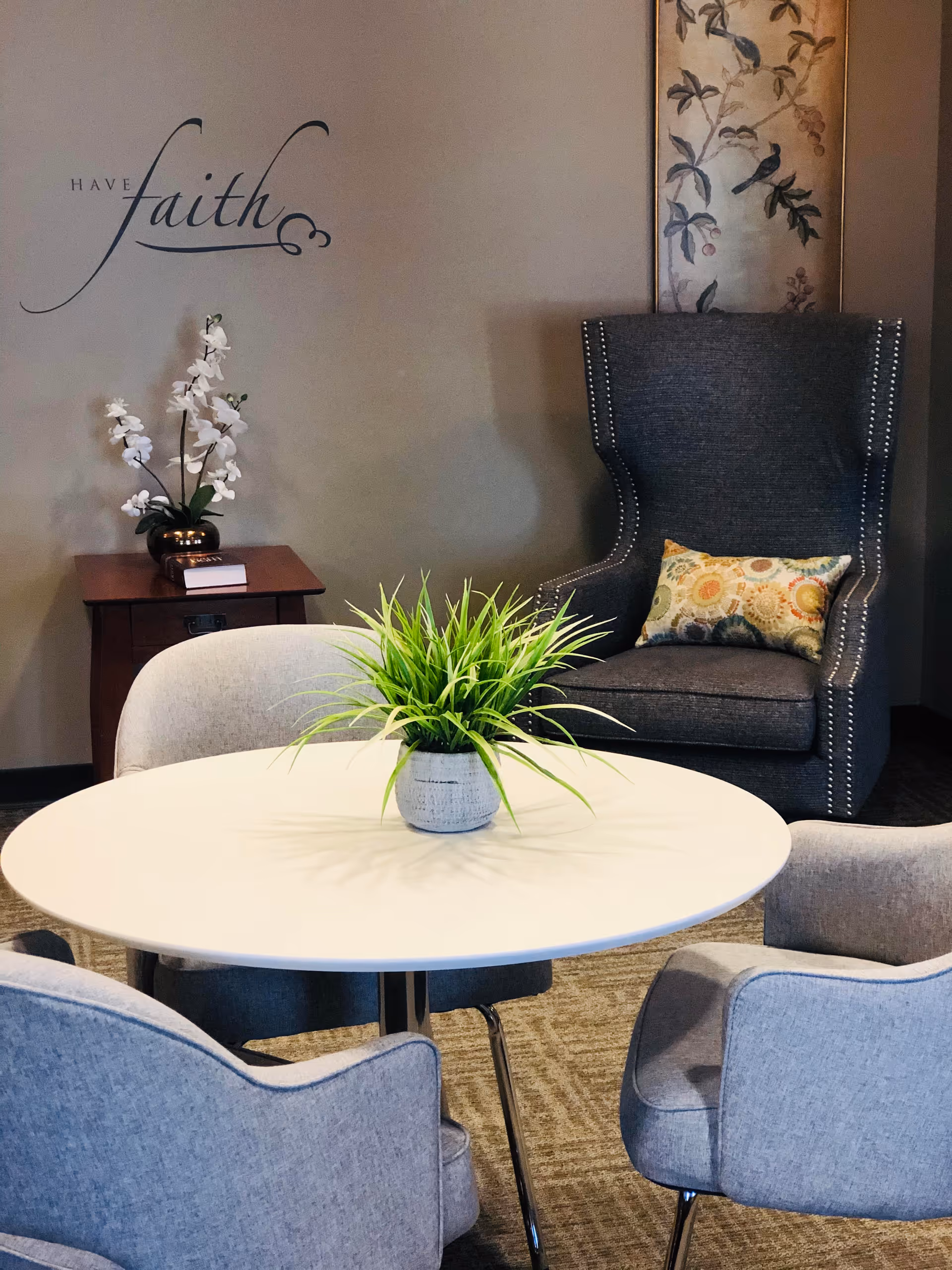 A cozy seating area with a round white table surrounded by three gray upholstered chairs. A potted green plant is centered on the table. In the background, there is a dark gray armchair with a decorative pillow, a small wooden side table with a white orchid plant and a book, and a wall decoration with the words 'Have faith' written on the wall.