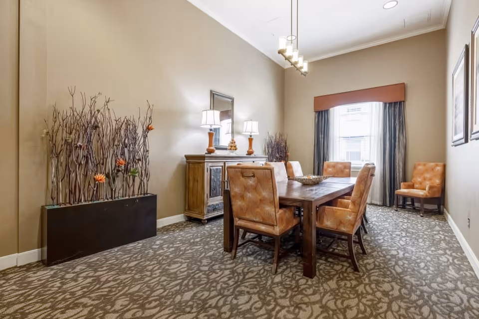 A well-lit dining room with a wooden dining table surrounded by six cushioned chairs. The room features patterned carpet flooring, beige walls, a window with curtains, a wooden sideboard with two lamps and a mirror above it, and decorative tall branches with flowers in a planter on the left side.