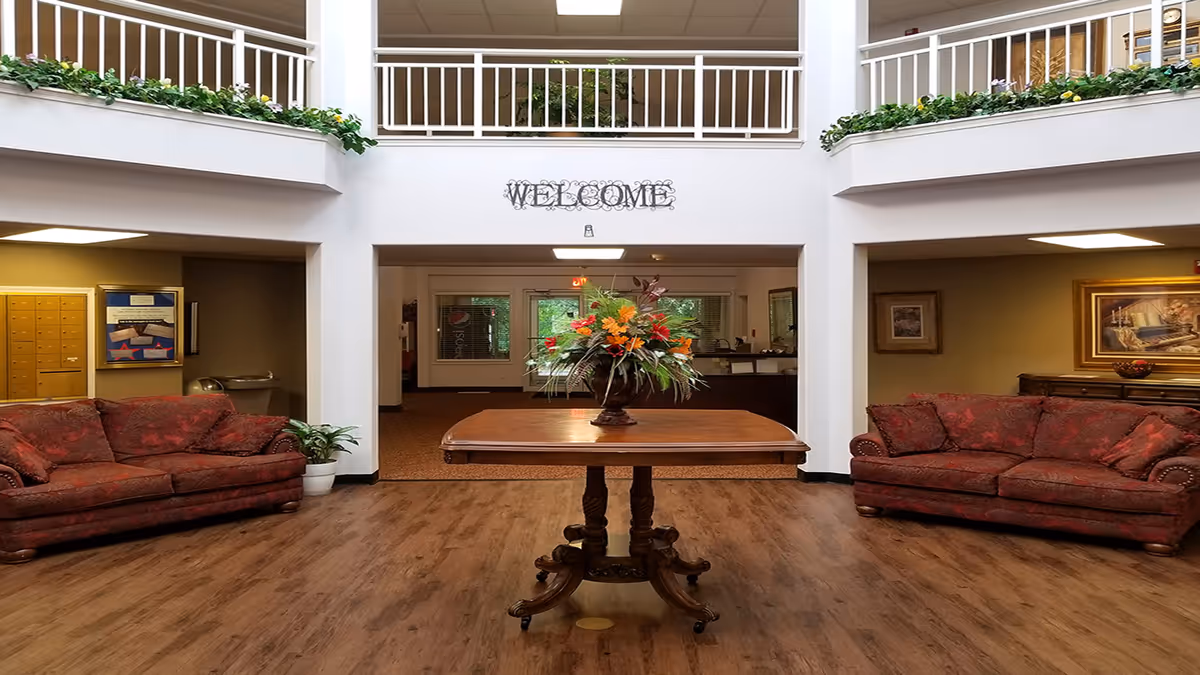 Interior view of a senior living facility lobby with a wooden table in the center holding a floral arrangement. Two red patterned sofas are placed on either side against the walls. Above the entrance is a decorative 'WELCOME' sign. The area has wooden flooring, white railings on the upper level, and some greenery along the balcony edges.