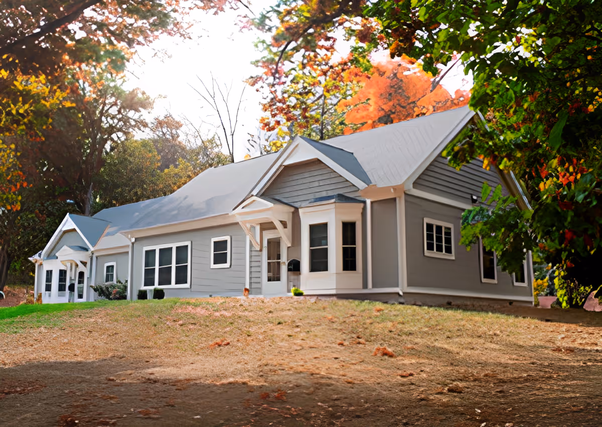 Exterior view of a single-story gray building with white trim, surrounded by trees with autumn foliage and a grassy area in front.
