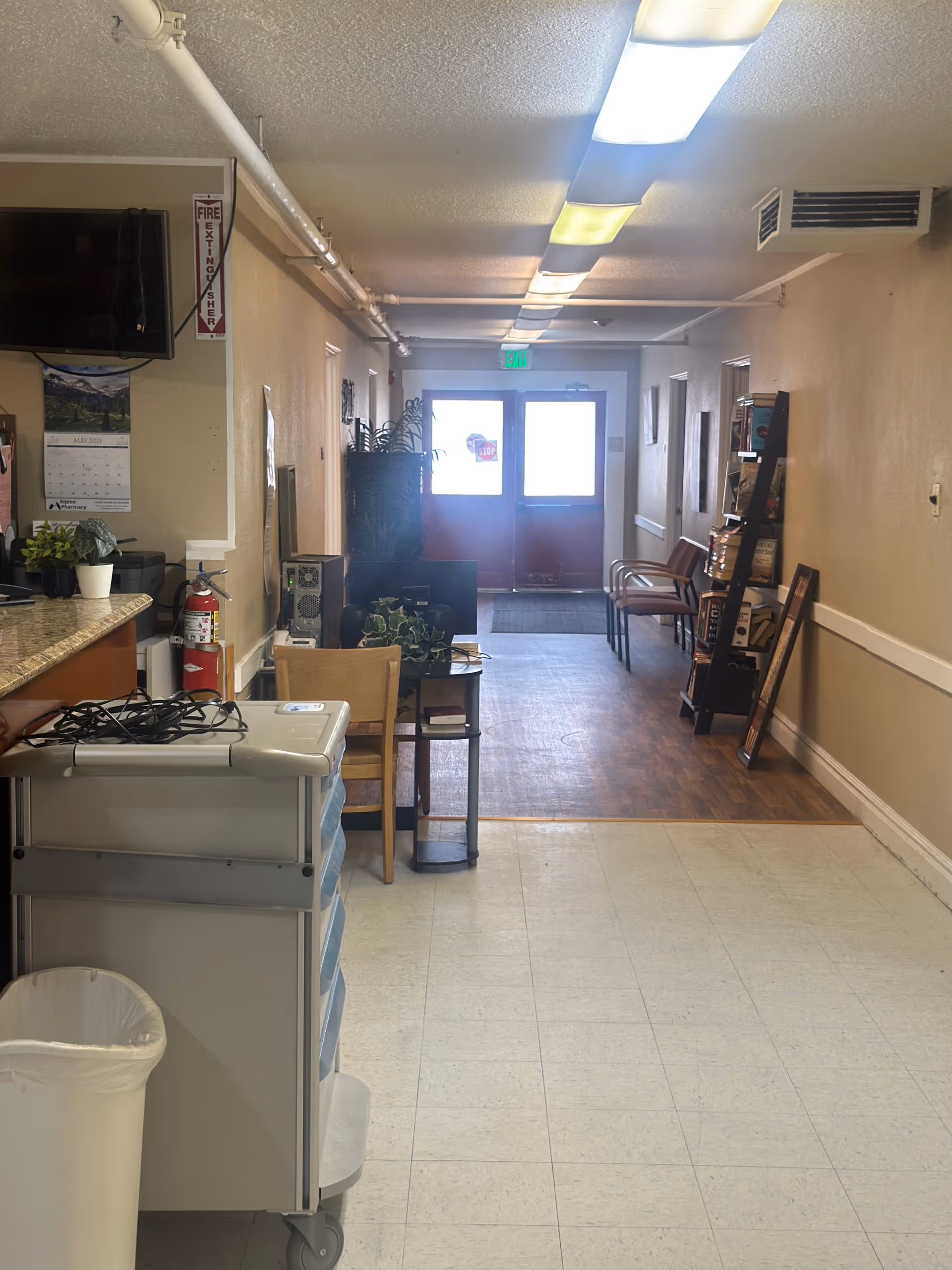 Interior hallway of an assisted living facility with a tiled floor transitioning to wood flooring near the exit doors. The hallway has beige walls, fluorescent ceiling lights, a fire extinguisher mounted on the wall, a calendar, a TV, a trash can, a small table with plants, chairs along the wall, and a bookshelf with various items. Double exit doors with glass panels are visible at the end of the hallway.