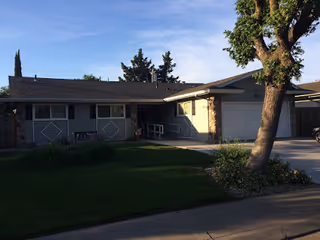 Single-story residential building with a front lawn, a tree, and a driveway leading to a garage under a clear blue sky.