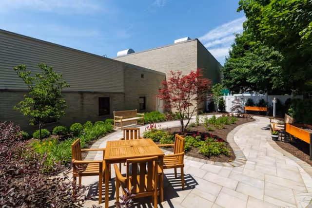 Outdoor patio area at LECOM - Parkside at North East featuring a paved walkway, wooden table with four chairs, several benches, landscaped garden beds with small trees and shrubs, and a clear blue sky.