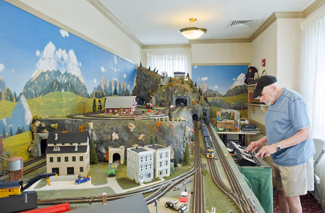 An elderly man operating a large model train set in a room with a scenic mountain mural on the walls. The train set includes miniature buildings, vehicles, tunnels, and tracks. The room is well-lit with natural light coming through sheer curtains.