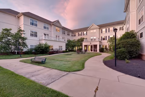 Outdoor courtyard area of a senior living facility with a paved walkway, green lawn, benches, trees, and a three-story building surrounding the courtyard under a pink and purple evening sky.