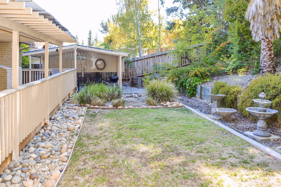 Outdoor garden area with a grassy lawn, decorative rocks along the edges, various shrubs and plants, a wooden fence in the background, a covered patio with a railing on the left, and a stone birdbath and fountain on the right.