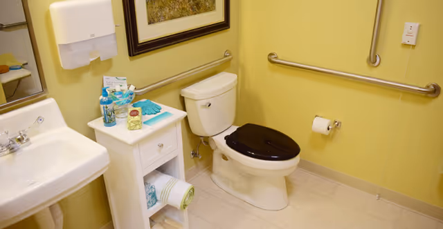 A clean bathroom with a white toilet featuring a black seat, a white sink with a silver faucet, a small white cabinet holding toiletries and a rolled towel, yellow walls with safety grab bars, a wall-mounted paper towel dispenser, and a framed picture above the toilet.