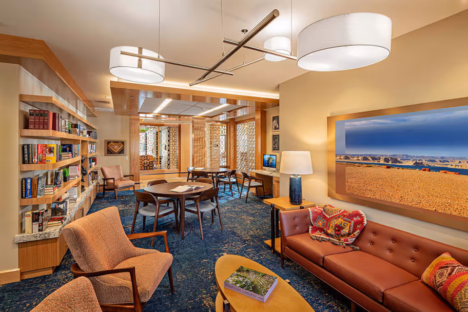 A cozy and well-lit common area in a senior living facility featuring a brown leather sofa with colorful patterned pillows, two upholstered armchairs, a wooden coffee table with a book, and a round table with chairs in the background. The room has a blue patterned carpet, wooden bookshelves filled with books, a desk with a computer, and large framed artwork on the wall. Modern ceiling lights illuminate the space.