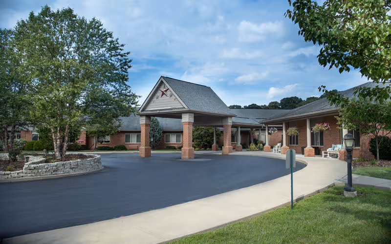 Exterior view of a senior living facility with a covered entrance supported by brick columns, a paved driveway, and landscaped greenery including trees and hanging flower baskets. The building is made of brick and has a gray shingled roof under a partly cloudy sky.