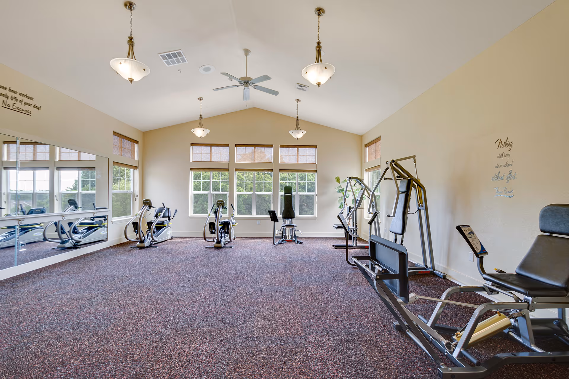 A bright exercise room with large windows letting in natural light. The room contains various exercise machines including stationary bikes and weight machines. The walls are light-colored, and there are ceiling fans and hanging light fixtures. A large mirror covers part of one wall, and motivational quotes are displayed on the walls.