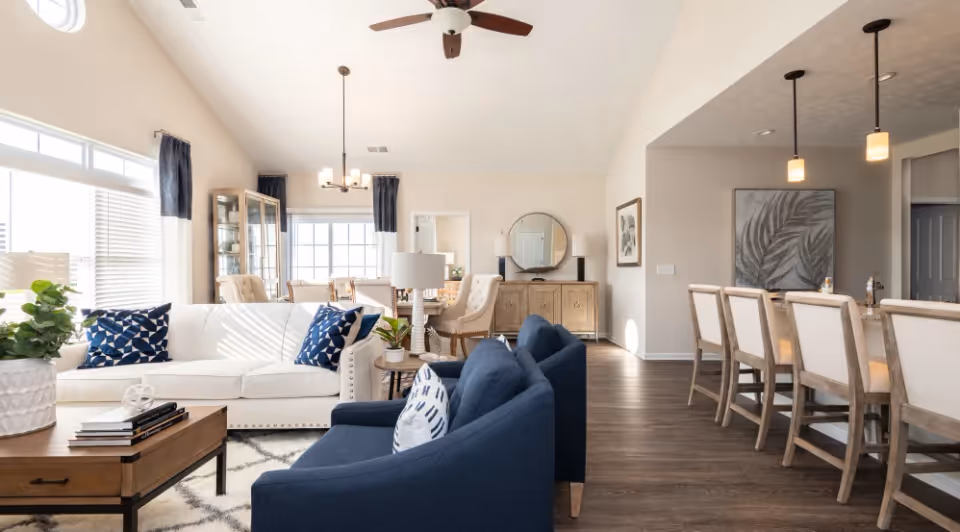 Bright and spacious living room with a white sofa and two blue armchairs arranged around a wooden coffee table. The room features large windows with white blinds and dark curtains, a ceiling fan, and a dining area with a wooden table and upholstered chairs in the background. To the right, there is a kitchen island with bar stools and pendant lights hanging above. The floor is a dark wood finish, and the walls are painted light beige.