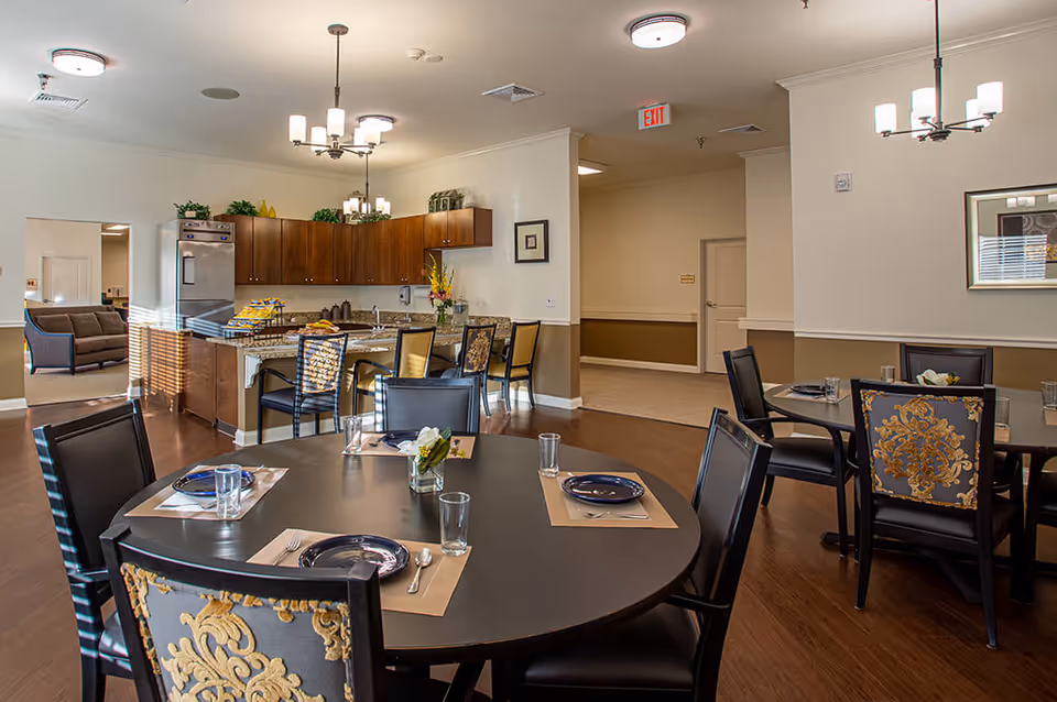 Dining area in Magnolia Springs Loveland featuring round tables set with placemats, plates, glasses, and silverware. The room has wooden flooring, decorative chairs with patterned upholstery, and overhead lighting fixtures. In the background, there is a kitchen area with wooden cabinets, a refrigerator, and a countertop with bar stools. A seating area with a sofa is visible through an open doorway.