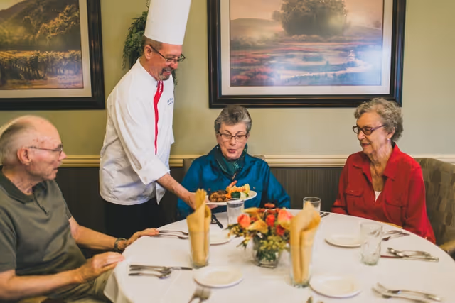 A chef serves plated food to three elderly residents seated around a round dining table in a dining room.
