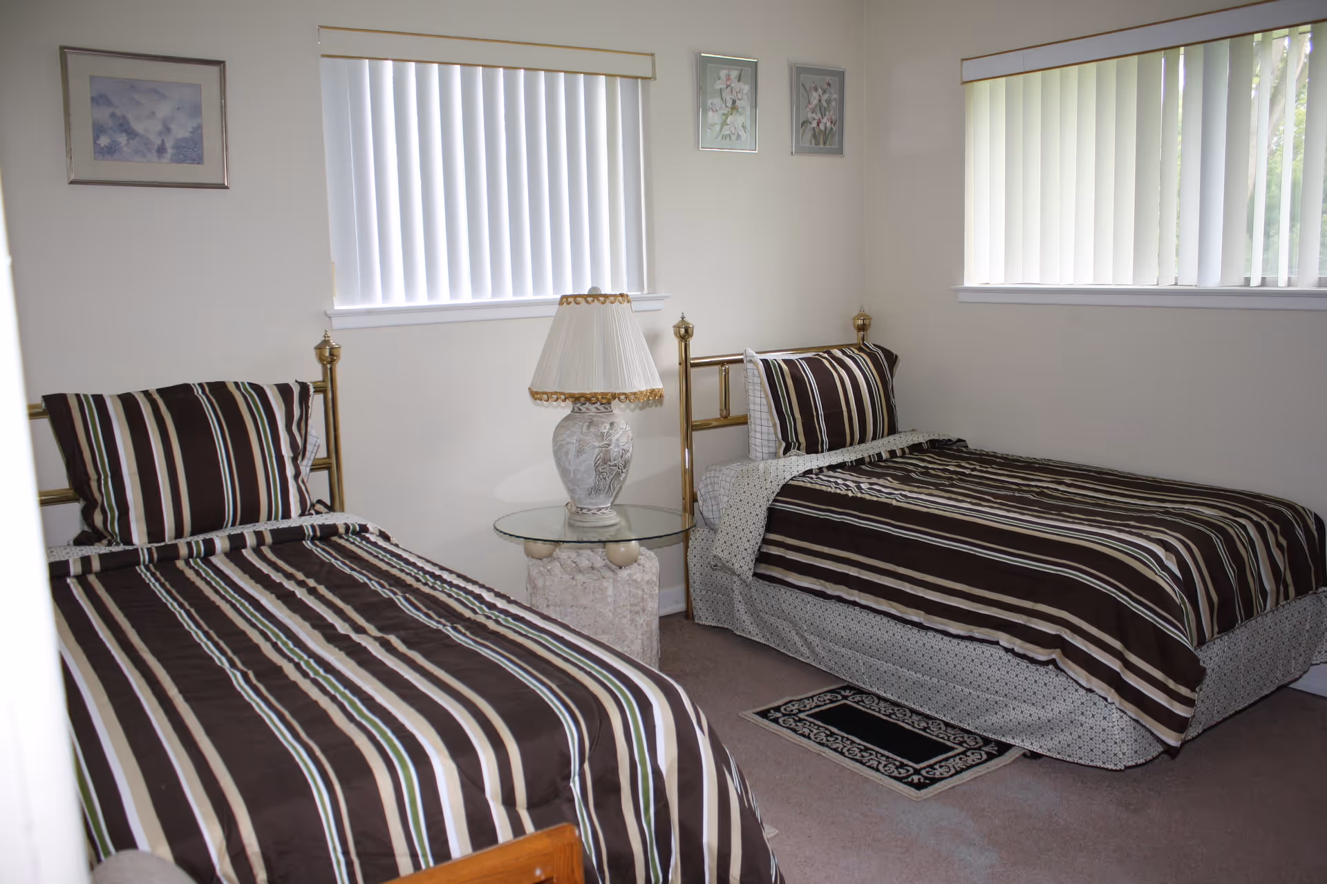 A bedroom with two single beds featuring brown, white, and beige striped bedding. Between the beds is a glass-top side table with a decorative lamp. The room has two windows with vertical blinds and three framed pictures on the walls.