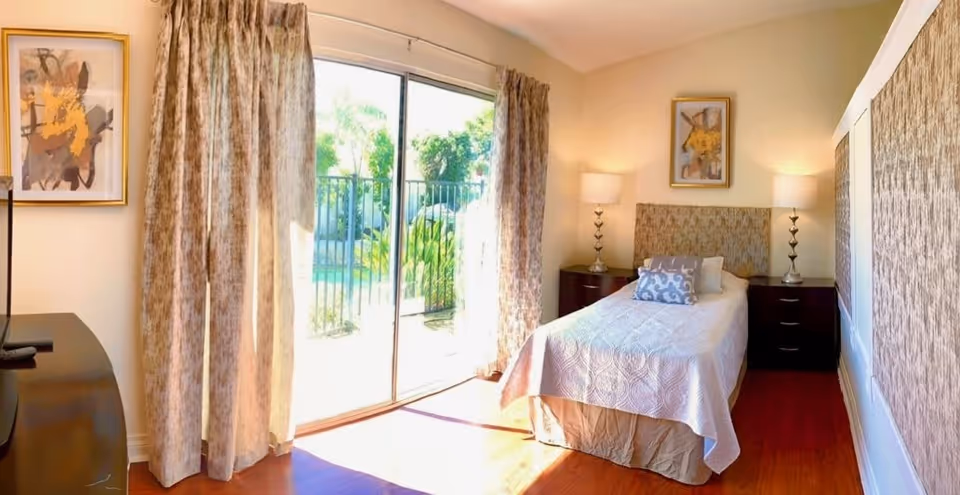 A bright bedroom with a single bed covered in a white quilt and decorative pillows. The bed is flanked by two dark wooden nightstands, each with a lamp. There are framed abstract paintings on the walls. A large sliding glass door with patterned curtains opens to an outdoor area with greenery and a pool.