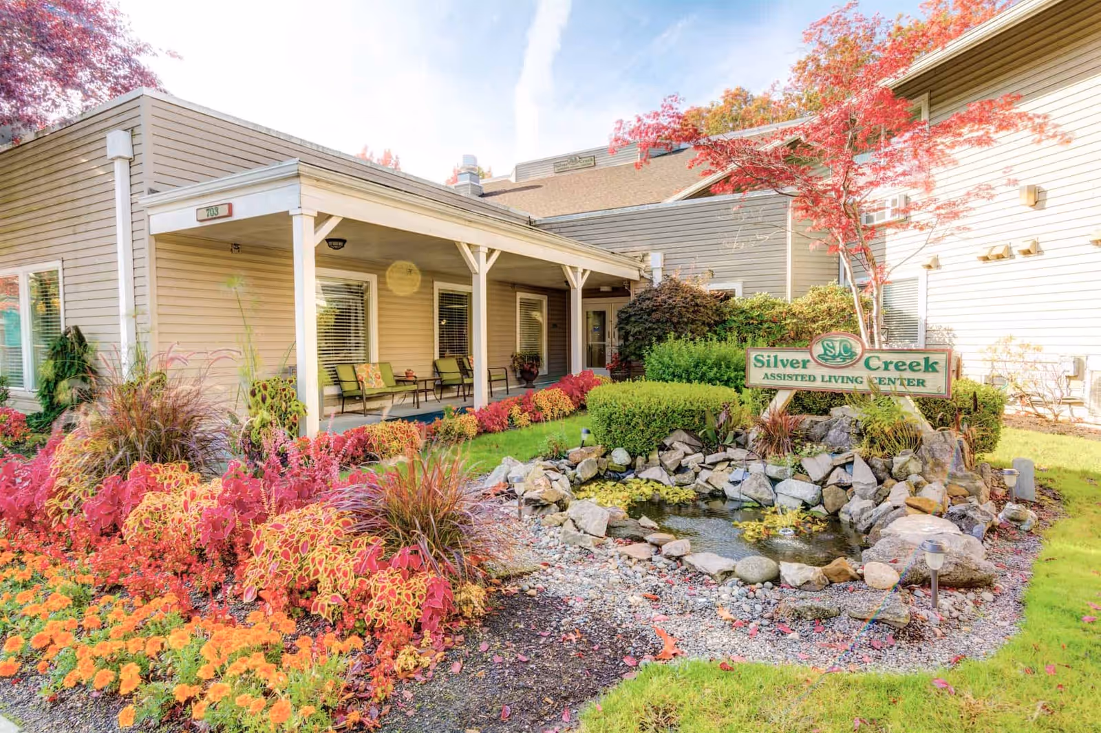 Exterior view of Silver Creek Assisted Living Center featuring a beige building with a covered porch, outdoor seating, colorful flower beds, a small pond with rocks, and a sign displaying the facility's name surrounded by greenery and trees with red leaves.