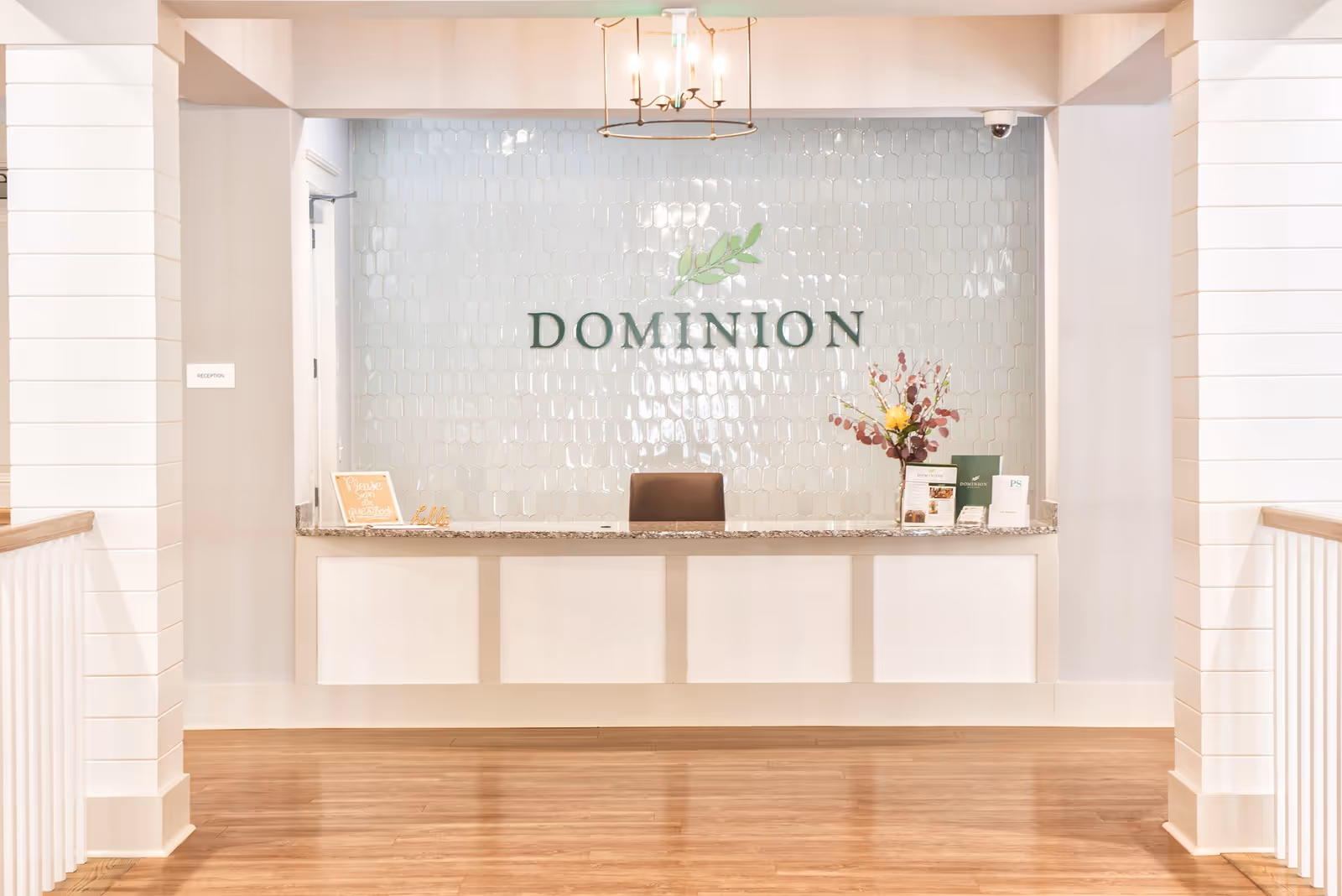 Reception desk area with a granite countertop and a chair behind it. The wall behind the desk features a decorative tile pattern with the word 'DOMINION' and a green leaf logo above it. There are informational brochures and a flower arrangement on the desk. The floor is wooden, and the surrounding walls are painted in light neutral colors.