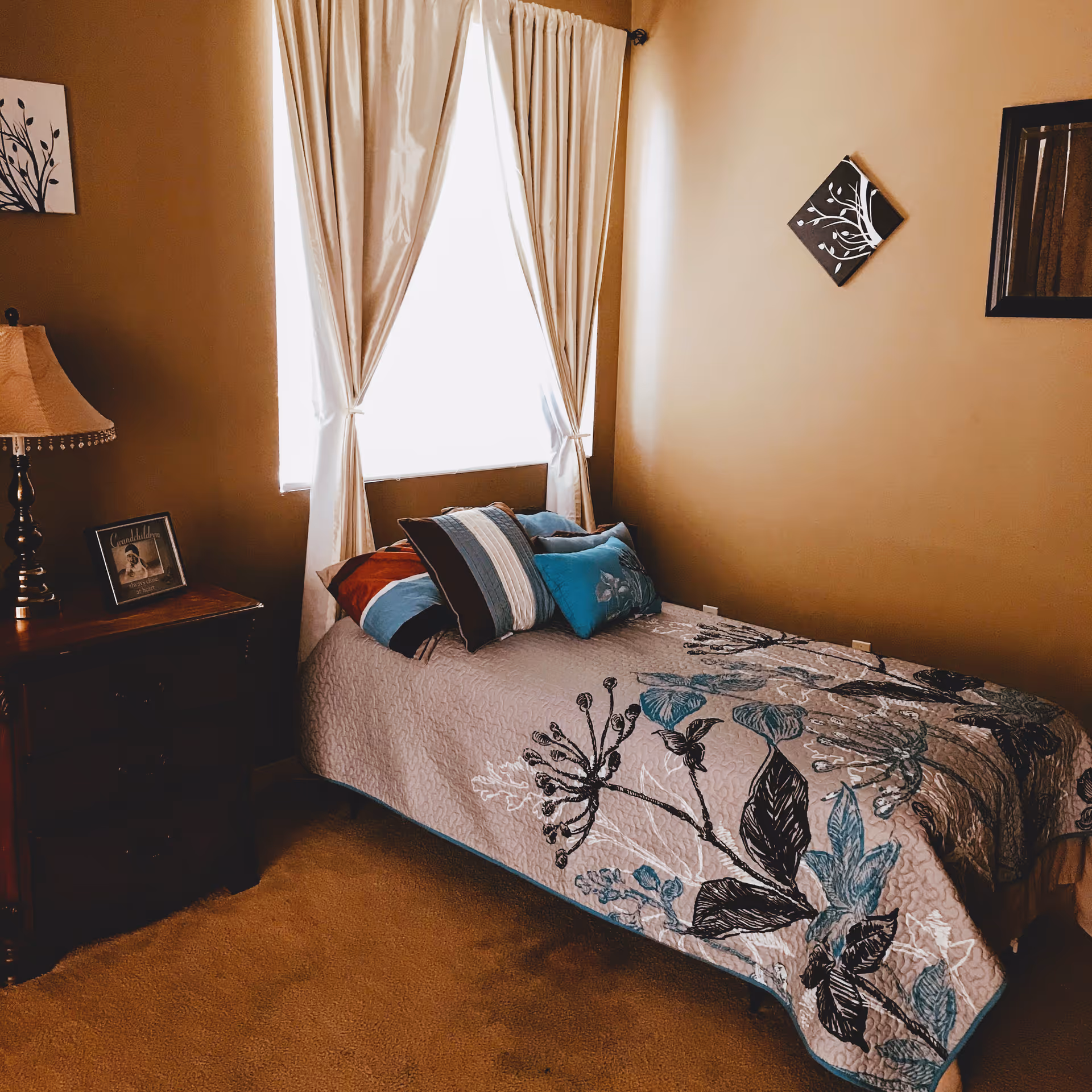 A cozy bedroom with a single bed covered in a floral-patterned quilt and several pillows. Next to the bed is a wooden nightstand with a lamp and a framed photo. The room has beige walls, a window with white curtains, and two pieces of wall art featuring tree designs.