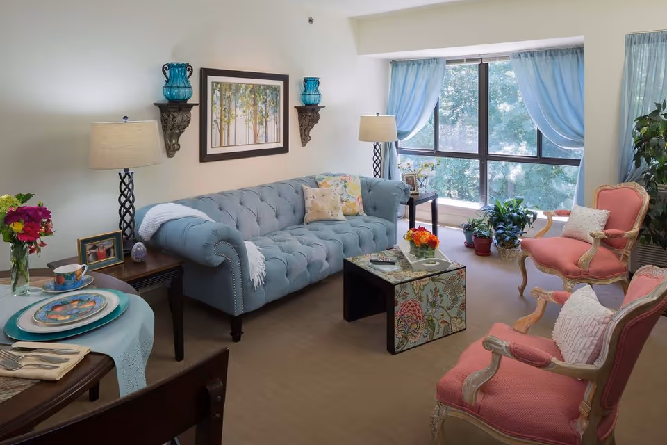 Bright living room with a tufted blue sofa, two pink armchairs, patterned coffee table and large window with blue curtains.