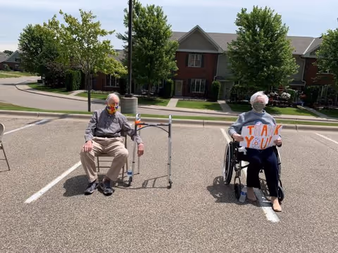 Two elderly individuals sitting in a parking lot with houses and trees in the background. One person is seated on a chair with a walker nearby, wearing a face mask. The other person is in a wheelchair holding a sign that says 'THANK YOU', also wearing a face mask.