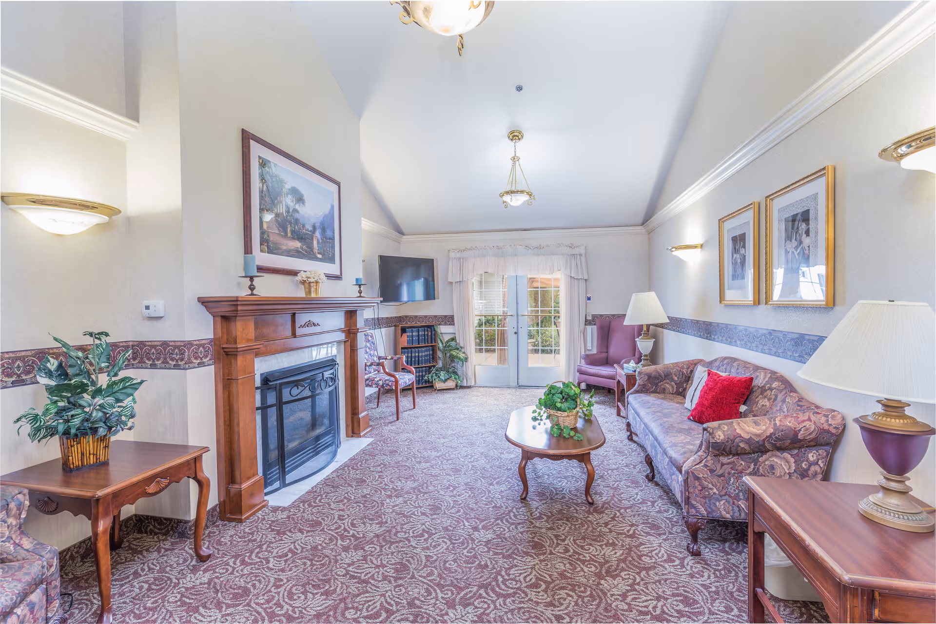 A cozy living room with a patterned carpet, a wooden fireplace with a painting above it, a flat-screen TV mounted on the wall, a floral-patterned sofa with a red pillow, two armchairs, wooden side tables with lamps, and a coffee table with a green plant. French doors with sheer curtains lead outside.