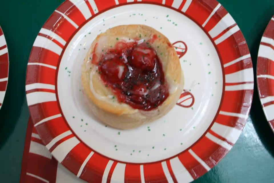 A close-up of a small pastry topped with red cherry filling and white icing, served on a red and white striped paper plate.