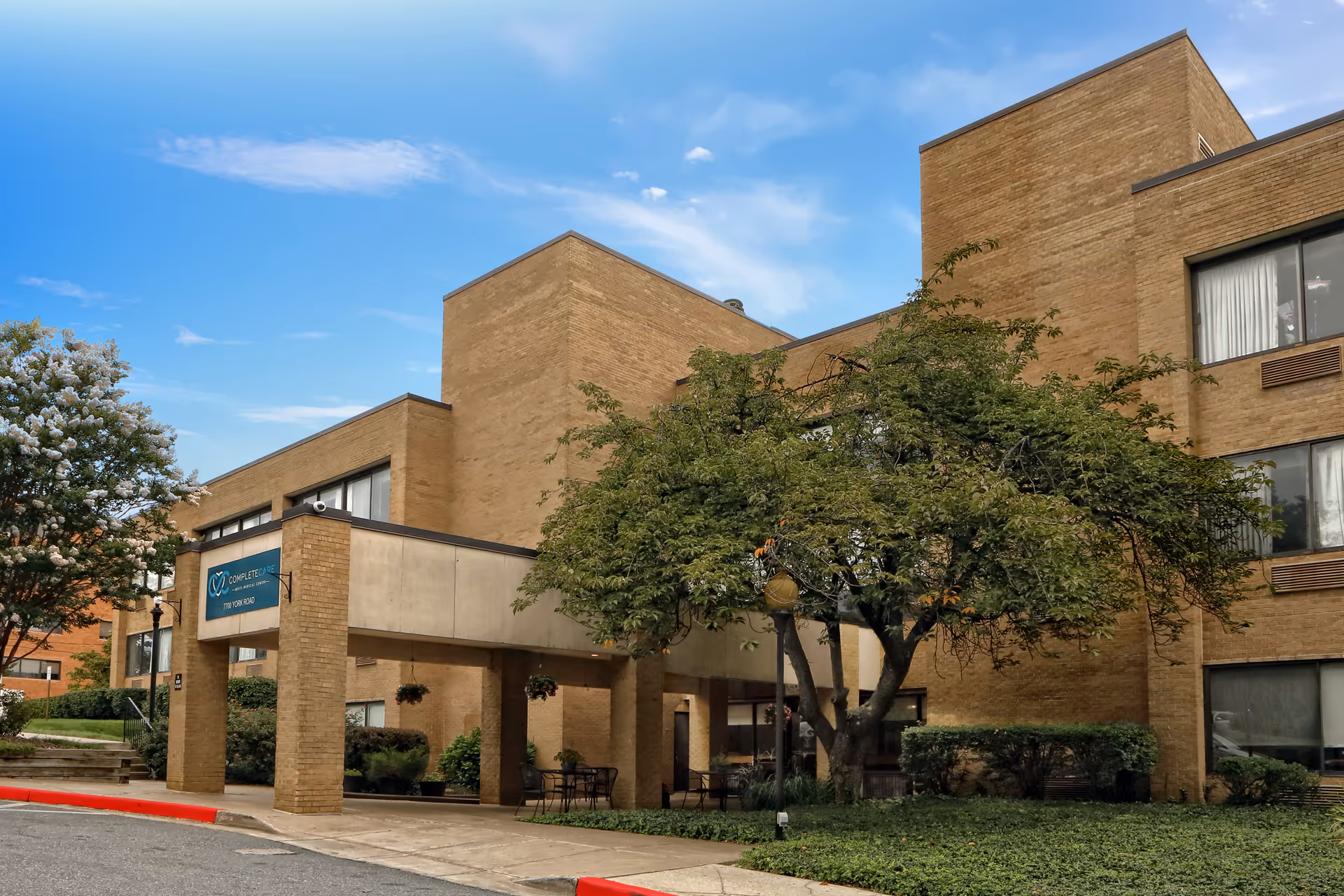 Exterior view of a multi-story brick building with a covered entrance and a sign that reads 'Complete Care at Multi-Medical Center'. There are trees and shrubs around the entrance area under a partly cloudy blue sky.