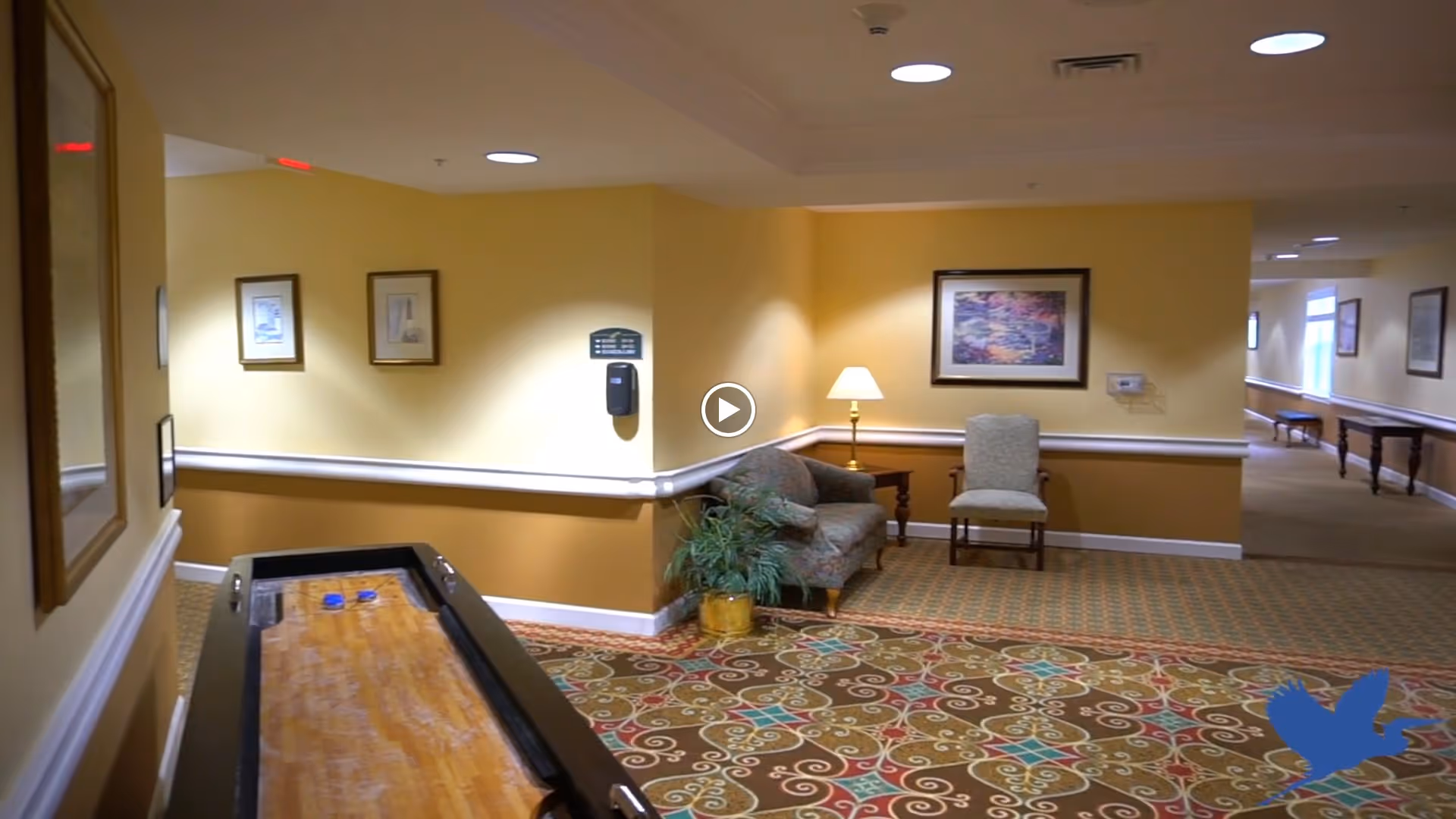 Interior hallway area with patterned carpet, yellow walls, framed artwork, a small seating area with a chair and a couch, a side table with a lamp, and a shuffleboard table in the foreground.