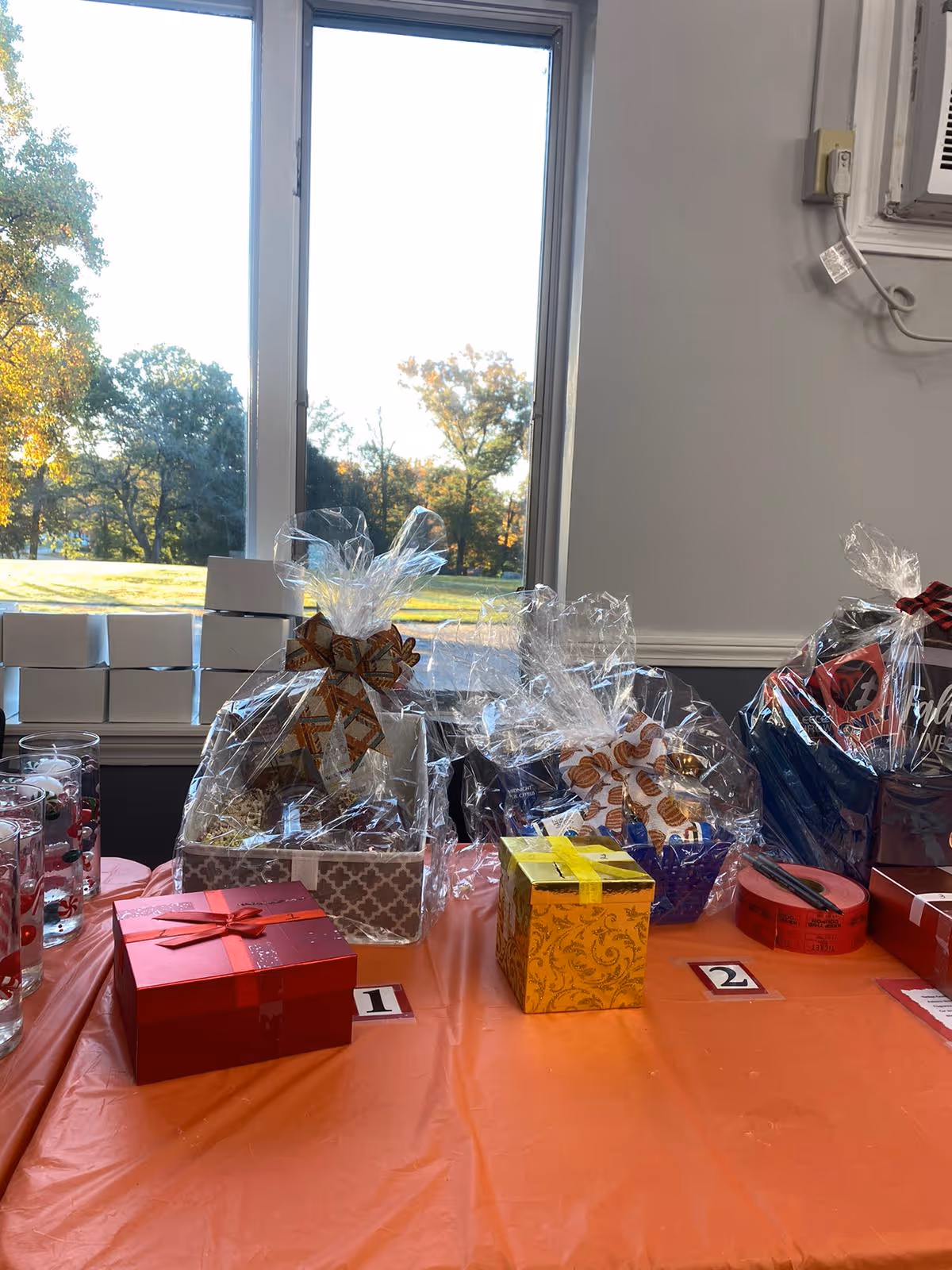 A table covered with an orange tablecloth displaying several gift baskets and wrapped boxes, each labeled with numbers 1 and 2. The table is set in front of a window showing a sunny outdoor scene with trees and grass.