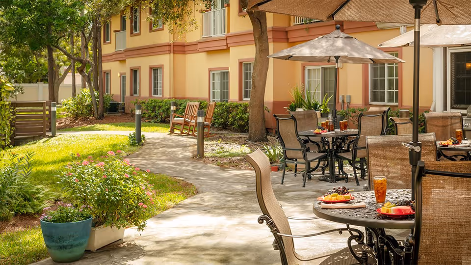 Outdoor patio area at Wickshire Tamarac with round tables and chairs under large umbrellas. Plates of fresh fruit and glasses of iced tea are set on the tables. The patio is adjacent to a yellow and red building with windows, surrounded by green grass, plants, and trees. A paved walkway leads through the garden area with wooden rocking chairs along the path.