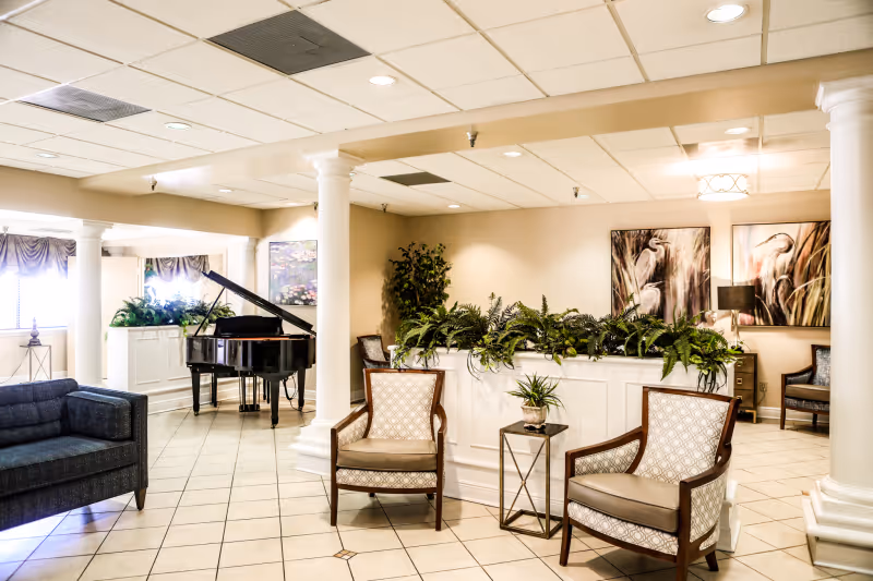 A bright and spacious living area in a senior living facility featuring a black grand piano, two upholstered armchairs with wooden frames, a dark blue sofa, several green plants on a white partition, and two large paintings of birds on the wall. The room has tiled flooring, white columns, and a drop ceiling with recessed lighting.