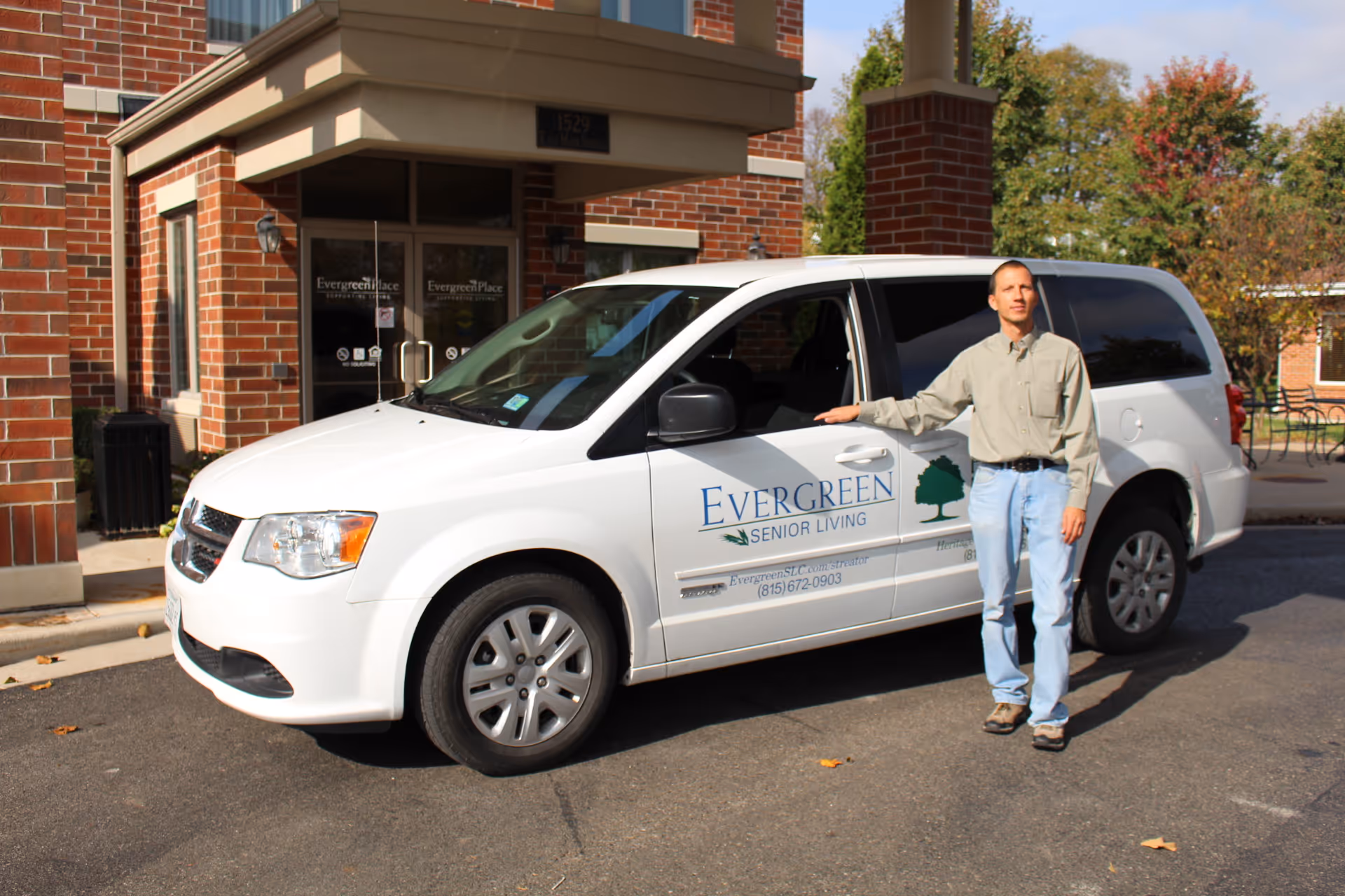 A man stands next to a white van with Evergreen Senior Living branding parked in front of a brick building entrance with glass doors. Trees with autumn foliage are visible in the background.