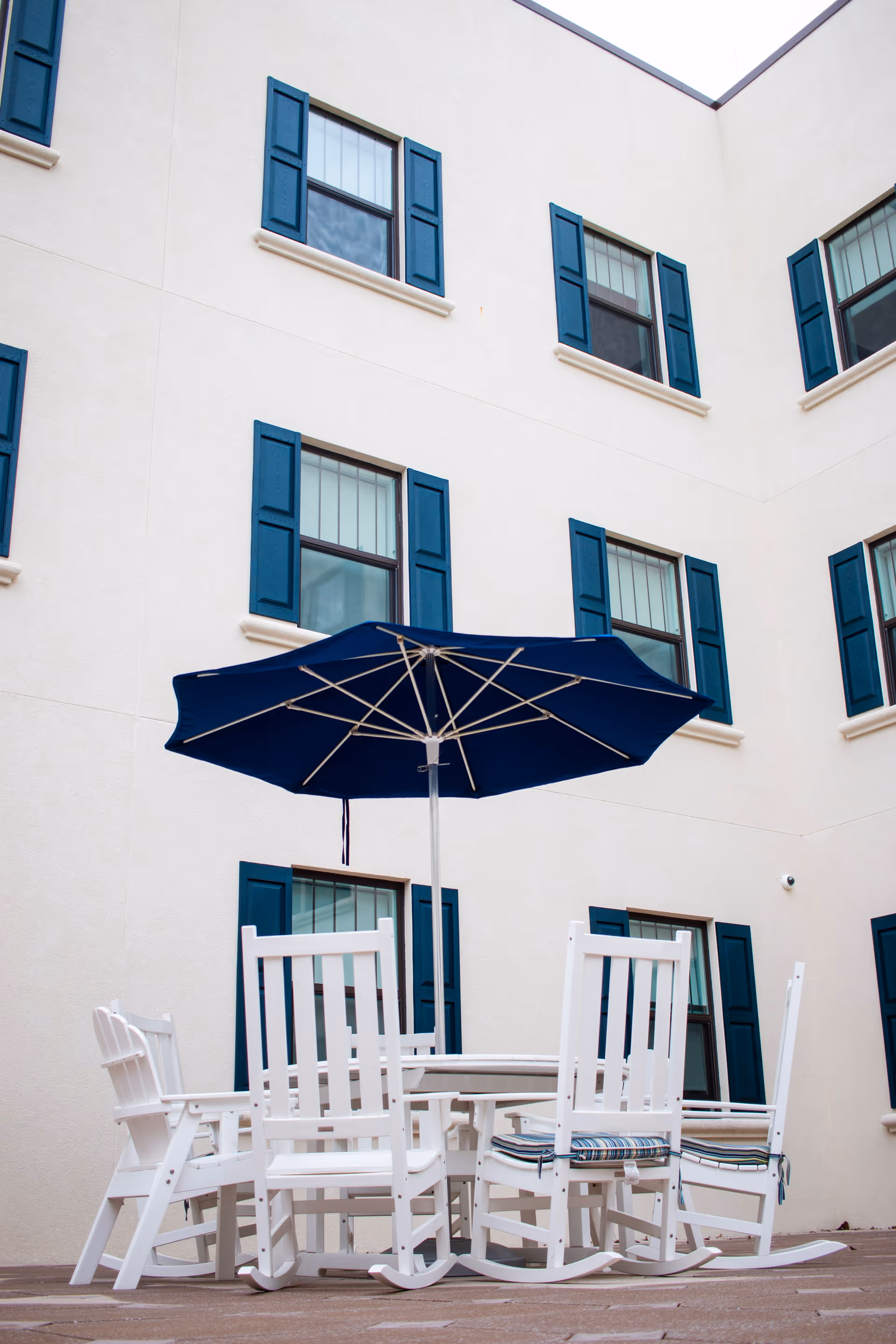 Outdoor courtyard area with white rocking chairs arranged around a round table with a large blue umbrella. The surrounding building has cream-colored walls and multiple windows with blue shutters.