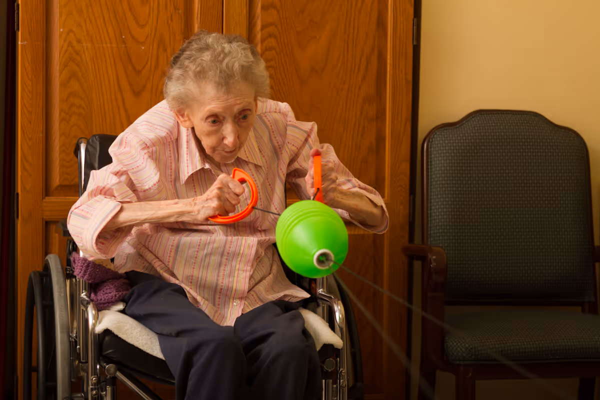 An elderly woman in a wheelchair is playing with a green and orange toy that involves pulling a string. She is indoors, sitting in front of a wooden door, with a green upholstered chair beside her.