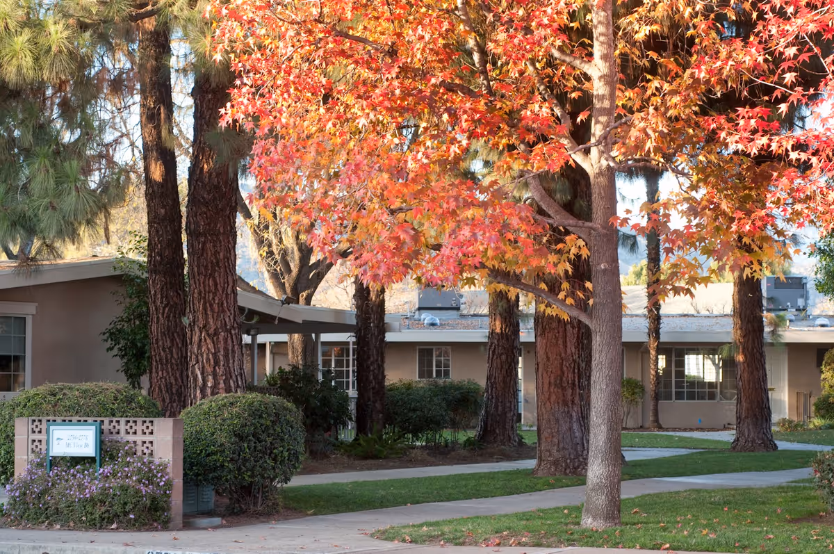 Outdoor view of a senior living facility named Hillcrest with a pathway, green grass, bushes, tall trees including one with vibrant red and orange autumn leaves, and a beige building in the background.