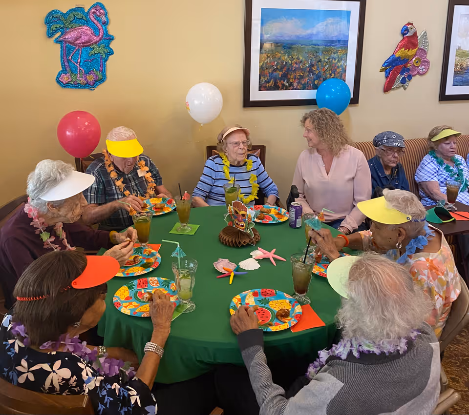 A group of elderly people and a caregiver sitting around a round table covered with a green tablecloth, enjoying a meal together. The table is decorated with colorful plates, drinks, and tropical-themed decorations. The people are wearing colorful visors and leis, and there are balloons and wall decorations including a flamingo and a parrot in the background.